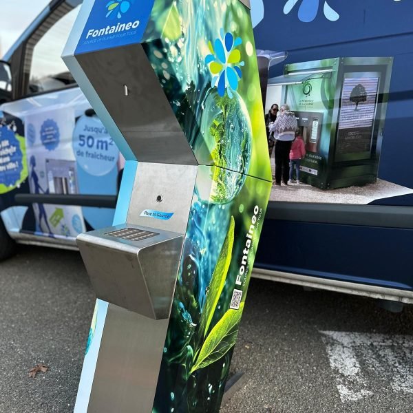Un distributeur d'eau moderne et de grande taille, aux motifs verts et bleus, est placé devant une camionnette. La machine est siglée « Fontaineo » et possède une base argentée. La camionnette en arrière-plan porte une marque similaire et des images de personnes utilisant la machine.