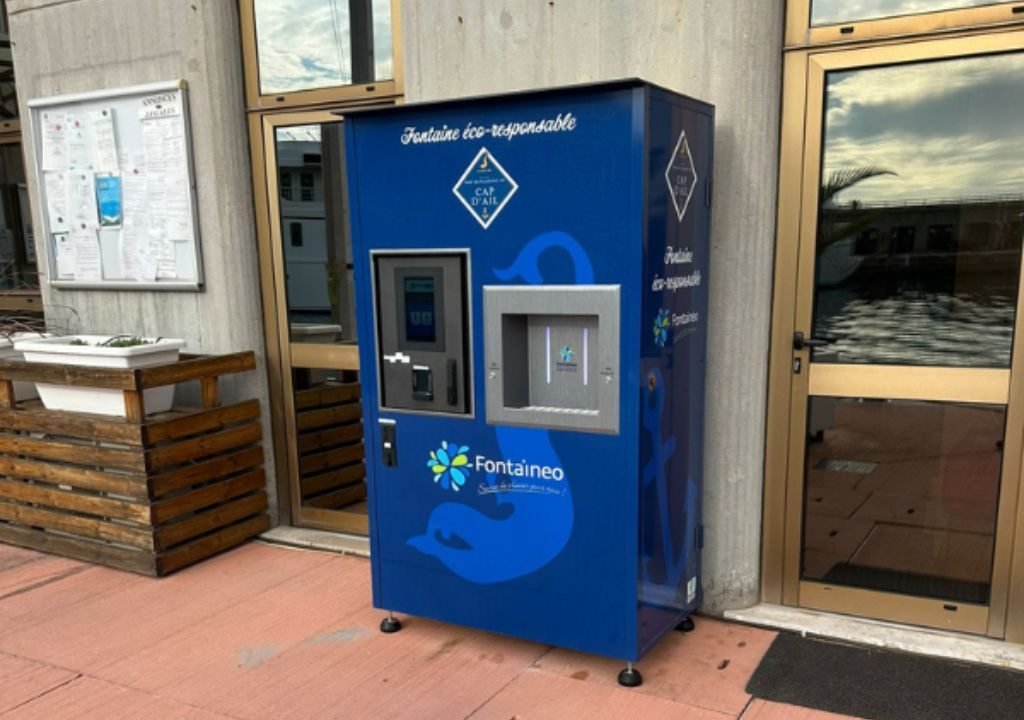 Un distributeur d'eau Fontaineo bleu est installé à l'extérieur d'un bâtiment, à côté de portes vitrées et d'un tableau d'affichage. La machine porte la mention « Fontaine éco-responsable » et permet de se servir de l'eau fraîche et de l'eau gazeuse.