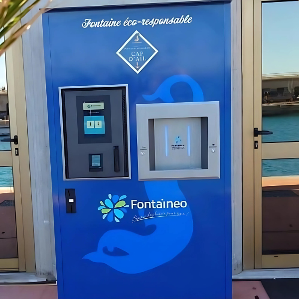 Station de remplissage d'eau bleue avec écran numérique et zone de distribution. Elle arbore des logos et des textes en français, promouvant l'éco-responsabilité. La station est située en extérieur, derrière des portes vitrées, avec vue sur l'eau et la marina en arrière-plan.