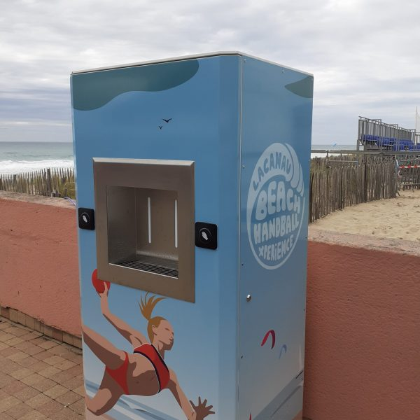 Un distributeur automatique bleu avec une petite fenêtre de distribution se dresse devant une plage en arrière-plan. Sur le côté, on peut voir une illustration d'une femme jouant au handball et l'inscription « Lacanau Beach Handball Experience ». Le sable et les vagues de l'océan sont visibles en arrière-plan.