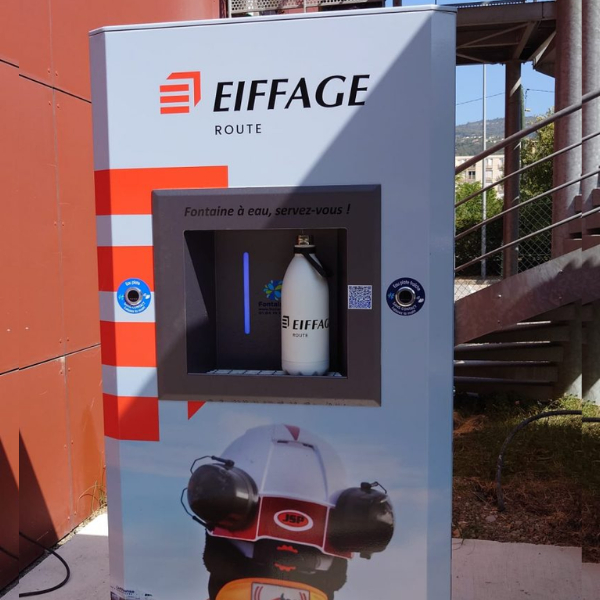 Un distributeur d'eau portant l'inscription « Eiffage Route » est installé à l'extérieur. Une bouteille blanche est visible à l'intérieur. En arrière-plan, un mur d'immeuble rouge, un escalier et une clôture. L'inscription « Fontaine à eau, servez-vous !.