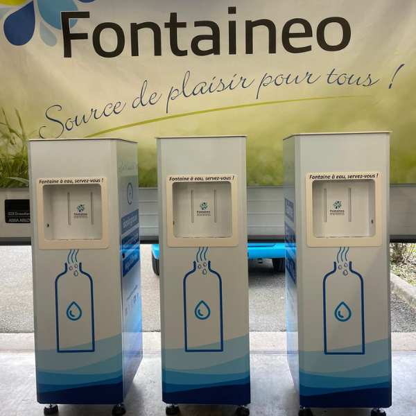 Trois distributeurs d'eau sont alignés devant une bannière sur laquelle est inscrit « Fontaineo » et un slogan en français. Chaque machine est blanche avec des touches de bleu, affichant une icône de bouteille d'eau et la phrase « Fonteaineo d'eau, servez-vous ! ».
