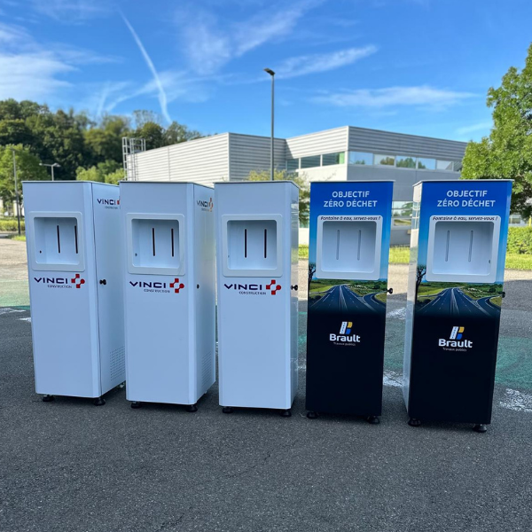 Cinq conteneurs à déchets sont installés à l'extérieur, chacun doté d'une ouverture rectangulaire. Les trois premiers arborent le logo « VINCI », tandis que les deux derniers affichent « Brault » et « Objectif Zéro Déchet ». Un bâtiment, des arbres et un ciel dégagé apparaissent en arrière-plan.