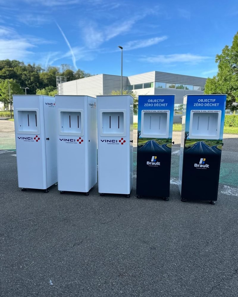 sourceo-light-fontaines-a-eau-reseau-fontaineo Une rangée de cinq bacs de recyclage extérieurs est installée sur une zone pavée. Les logos « VINCI » et « Brault » sont apposés sur les bacs, à côté des mots « Objectif Zéro Déchet ». Des arbres et un bâtiment sont visibles à l'arrière-plan.