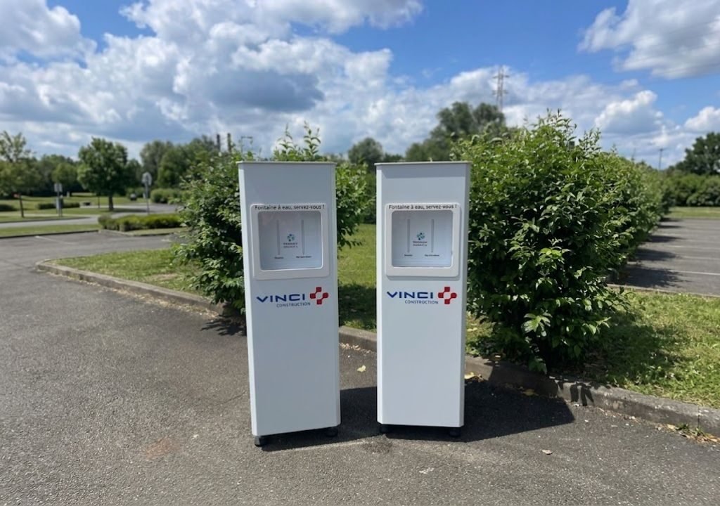 Deux distributeurs de gel hydroalcoolique blancs portant la marque VINCI sont placés côte à côte sur une surface asphaltée, dans un espace extérieur agrémenté de buissons verts, sous un ciel partiellement nuageux, avec une fontaine à eau de chantier visible à proximité, en arrière-plan.